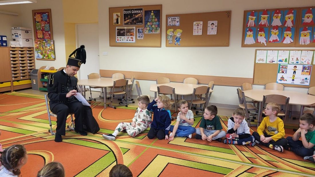 Siedzący górnik w stroju galowym rozmawiający z siędzącymi w siadzie tureckim dzieciakami z przedszkola