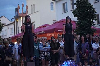 dwie aktorki w mrocznych, czarnych strojach stoją nieruchomo na podwyższeniach przed publicznością; jedna z nich trzyma duży trójząb
