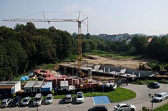 Widok z góry na plac budowy z wykopem pod fundamenty, strefą ze składowanymi materiałami, żurawiem wieżowym i pobliskim parkingiem