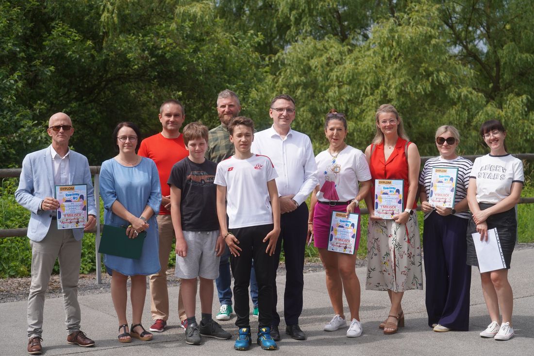 Grupa laureatów i organizatorów stoi na zewnątrz w rzędzie, pozując do zdjęcia. Niektóre osoby trzymają w rękach dyplomy lub foldery z dokumentami