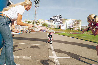 Kobieta macha flagą przed małym dzieckiem jadącym na rowerku w stronę mety