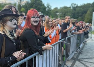 16. Grupka dziewczyn, jedne siedzą drugie stoją pozuje do zdjęcia. W tle widać innych siedzących ludzi na ławkach.