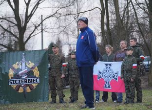Organizator uroczystości na tle dzieci i dorosłych w żołnierskich strojach trzymających transparenty z polskimi symbolami