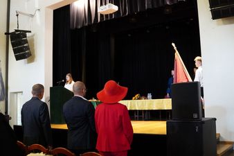Uczestnicy uroczystości, w tym kobieta w czerwonym kapeluszu, stoją tyłem do obiektywu i obserwują oficjalną część wydarzenia na scenie.