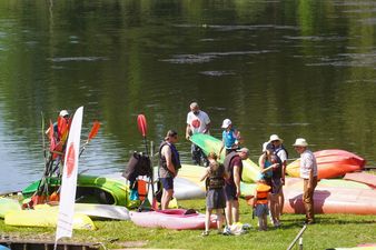 Grupa osób na trawiastym brzegu rzeki, przygotowująca kajaki do spływu, z wiosłami i kamizelkami ratunkowymi.