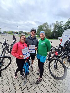 Trzy uśmiechnięte osoby w strojach sportowych stoją z rowerami na placu i prezentują otrzymane dyplomy