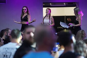 Trzy tancerki występujące na scenie w fioletowym oświetleniu podczas imprezy. Jedna trzyma wachlarz, druga ma warkocze i malunki na twarzy