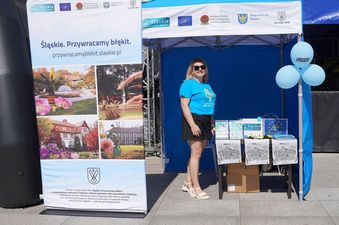 Kobieta w niebieskiej bluzce stojąca przy stoisku z gadżetami, obok baner ze zdjęciami i napisem Śląskie. Przywracamy błękit