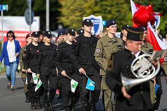 Młodzież z klas mundurowych maszerująca w czarnych i oliwkowych mundurach taktycznych z małymi flagami w dłoniach, mijająca muzyka z waltornią