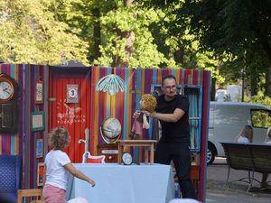 Aktor na tle kolorowej, pasiastej scenografii teatrzyku plenerowego animuje kukiełkę chłopca przed małym dzieckiem