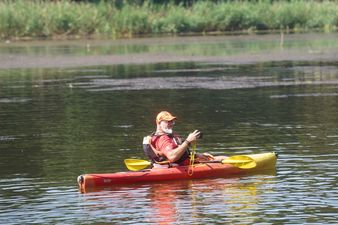 Mężczyzna w pomarańczowej czapce i czerwonej koszulce, siedząc w czerwonym kajaku, trzyma aparat fotograficzny i patrzy w niego