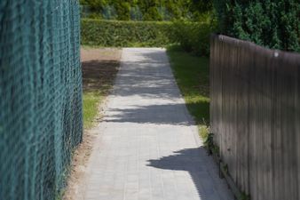 Wąska ścieżka z kostki brukowej prowadząca w głąb ogrodu po chodniku na ulicy Bananowej. Po lewej stronie znajduje się wysoki płot z siatki, a po prawej drewniane ogrodzenie. W tle widać gęste zielone krzewy i drzewa