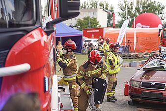 Strażacy wyciągają sprzęt z bocznej skrytki czerwonego samochodu gaśniczego, z przodu po prawej widać zniszczone czerwone auto z wybitą szybą
