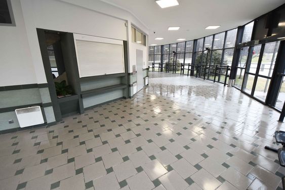 Przestronny, jasny hol z przeszkloną ścianą wychodzącą na zewnątrz. Na podłodze wzorzyste kafelki. Widoczne zamknięte okienko recepcyjne z białą roletą i grzejniki konwektorowe na ścianach