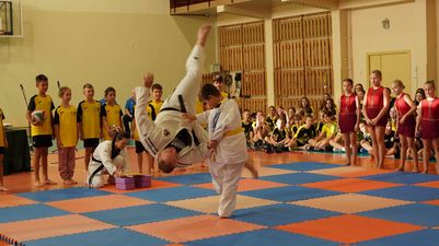 Pokaz judo w wykonaniu dwojga zawodników na macie; jeden z nich wykonuje rzut, pozostali uczniowie obserwują występ