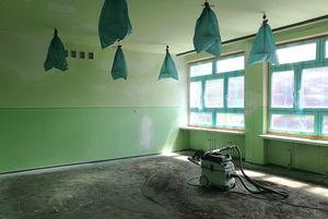Odnowiona sala pomalowana na intensywny zielony kolor. Lampy na suficie są zabezpieczone niebieską folią. Na środku brudnej podłogi stoi odkurzacz przemysłowy z rurą.
