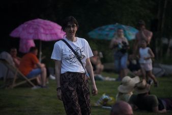 Kobieta w okularach, białej koszulce z kwiatowym wzorem i wzorzystych spodniach, stoi na trawie w wieczornym oświetleniu. W rozmytym tle widać innych ludzi i różowy parasol plażowy
