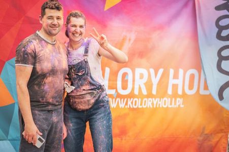 Uśmiechnięta kobieta i mężczyzna umazani proszkami holi pozują na tle kolorowej ścianki z napisem „KOLORY HOLI”