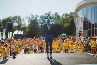 Kobieta w czarnej koszulce z podniesionymi ramionami stojąca na podium, w tle grupa ćwiczących dzieci