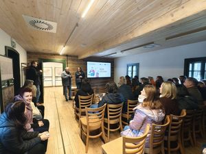 Ujęcie z boku uczestnicy spotkania siedzą na krzesłach na sali, patrzą na ekran gdzie wyświetla się slajd z napisem śląsk cieszyński