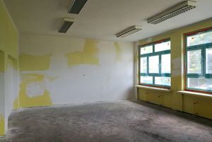 Pusta, remontowana sala z dużymi oknami. Ściany są przygotowane do malowania z żółtymi przetarciami. Na suficie widoczne podłużne lampy, a podłoga jest surowa i zapylona