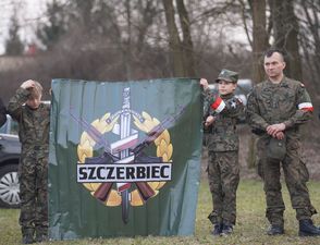 Mężczyzna oraz chłopcy w wojskowych strojach trzymający sztandar z napisem Szczerbiec