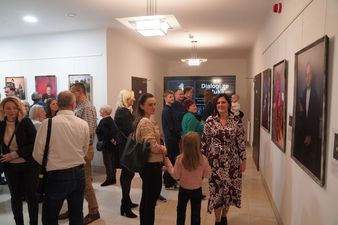 Tłum ludzi na otwarciu wystawy sztuki; z boku widoczne portrety i tablica informacyjna