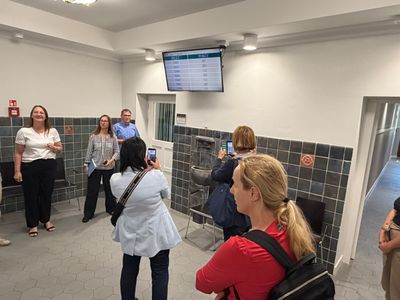 Grupa osób w urzędzie przygląda się ekranowi systemu kolejkowego z napisami „BILET” i „POKÓJ”, a dwie kobiety robią mu zdjęcia smartfonami. W tle stoją dwaj pracownicy