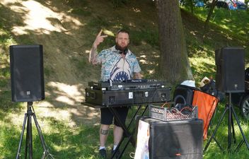 Mężczyzna didżej w koszuli z motywem rekinów stoi za konsolą Pioneer DJ, ustawioną plenerowo pod drzewami