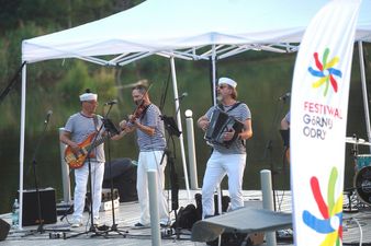 Pionowa flaga z logo Festiwalu Górnej Odry po prawej stronie kadru. W tle na pomoście wokalista z akordeonem, skrzypek oraz gitarzysta basowy w strojach marynarskich