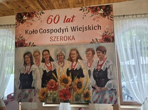 Plakat z napisem „60 lat Koło Gospodyń Wiejskich Szeroka” oraz z namalowanymi postaciami w strojach ludowych KGW