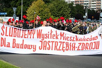 Uczestnicy pochodu, w tym górnicy w galowych mundurach z pióropuszami i orkiestra, niosący szeroki transparent z czerwonym napisem "ŚLUBUJEMY Wysiłek Mieszkańców Dla Budowy JASTRZĘBIA-ZDROJU"