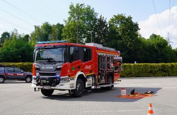 Czerwony samochód strażacki marki Scania zaparkowany na placu z otwartą boczną skrytką na sprzęt ratowniczy. Obok na macie leżą narzędzia hydrauliczne