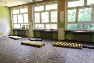 Remont sali lekcyjnej. Na podłodze leżą zdemontowane żebrowe grzejniki. Ściany są częściowo odrapane, pomalowane na zielono i biało. Na parapecie pod dużym oknem stoi godło Polski