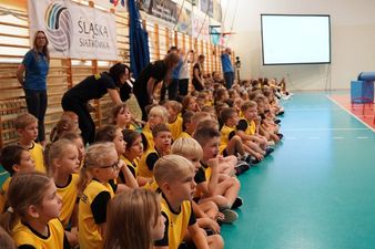 Grupa dzieci klas 1-4 ubranych w stroje siatkarskie, siedząca za boiskiem przy drabinkach