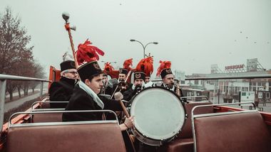 Zdjęcie przedstawia orkiestrę w otwartym autobusie piętrowym, widok na Supersam.