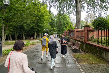 Przewodniczka i turyści spacerują kamienną alejką w gęsto zalesionym parku. Po prawej stronie ciągnie się długi, murowany płot z metalowym ogrodzeniem oraz stoją drewniane ławki parkowe