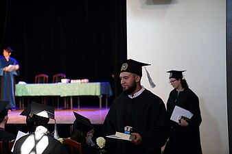 Uśmiechnięty absolwent w birecie i todze schodzi ze sceny, trzymając w ręku białą różę oraz pamiątkową statuetkę na książce