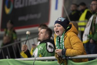 Młody kibic w żółtej kurtce i niebieskiej czapce głośno dopinguje z trybun, w tle inny mężczyzna trzyma pałkę do bębna