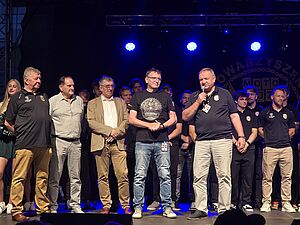 Sportowcy na scenie, jeden z nich przemawia do mikrofonu podczas wydarzenia plenerowego