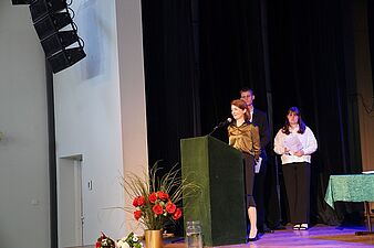 Opiekunka roku w eleganckiej satynowej bluzce przemawia do mikrofonu przy zielonej mównicy. Za nią na scenie stoją dwie inne osob
