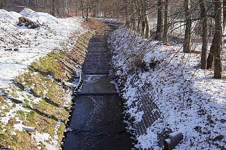 Uregulowane, wyłożone płytami koryto rzeki/strumienia płynące wzdłuż ośnieżonych, zadrzewionych brzegów