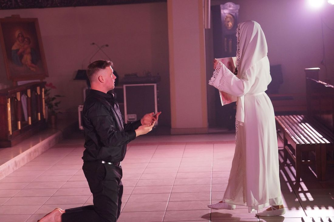 Ubrany na czarno mężczyzna klęczy na posadzce przed postacią w białych szatach