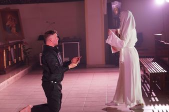 Klęczący aktor utożsamiający postać Księdza Machy, przed Nim stojąca kobieta ubrana na biało trzyma w rękach chustę z wizerunkiem Chrystusa