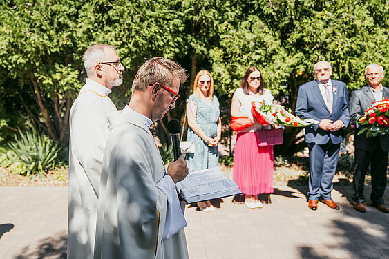 Ksiądz stoi na dworze, trzyma mikrofon i spogląda w pismo święte, przed nim stoją ludzie z kwiatami