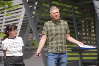 Wojciech Flak pracownik Urzędu Miasta w koszuli w kratę stoi na zewnątrz z otwartymi ramionami, trzymając w jednej ręce długopis, a w drugiej folder z dokumentami. Obok niego stoi kobieta w okularach