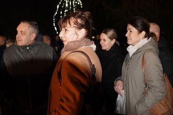 Kilka osób, w tym kobieta w okularach i brązowym płaszczu, uchwyconych z profilu podczas nocnego wydarzenia