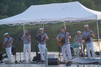 Sześciu muzyków zespołu szantowego w pasiatych koszulkach i marynarskich czapkach gra koncert na pływającym pomoście pod białym zadaszeniem