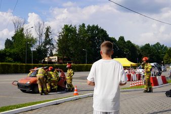 Chłopiec w białej koszulce tyłem do obiektywu przygląda się pracy grupy strażaków, którzy zabezpieczają uszkodzony, pomarańczowy samochód.