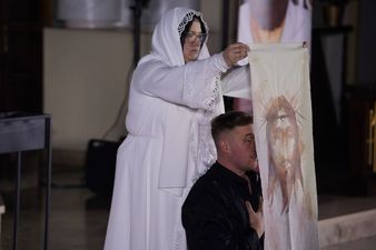Klęczący aktor utożsamiający postać Księdza Machy, za Nim stojąca kobieta ubrana na biało trzyma przed Nim w rękach chustę z wizerunkiem Chrystusa